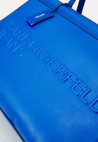 Sininen nahkainen kassi, jossa on kohokuvioitu "KARL LAGERFELD" -logo. Ominaisuuksina sileä pinta ja tukevat kahvat. Mukana sininen tagi.