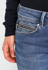 Primer plano de una mano al lado de unos jeans de mezclilla azules con un bolsillo para monedas con cremallera y una camisa negra de manga larga visible en la muñeca.