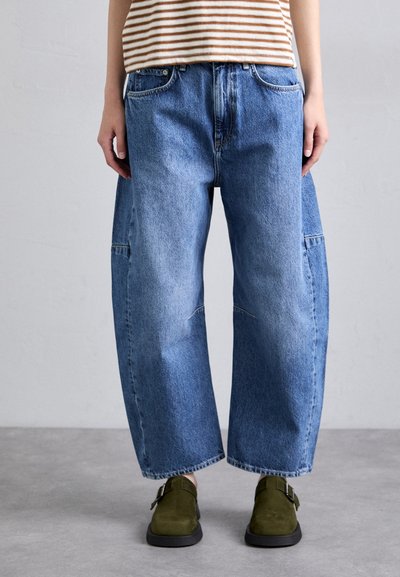 Jean en denim bleu ample avec une finition délavée, présentant une coupe décontractée et des coutures visibles. Portés avec des chaussures en daim vert foncé.
