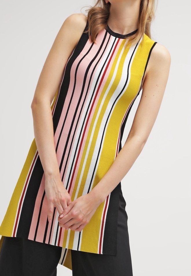 Tunique en tricot sans manches avec des rayures verticales rose, jaune, blanche, noire et rouge, portée sur un pantalon noir.