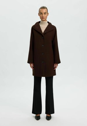 Femme debout portant un manteau boutonné marron foncé sur un col roulé beige, un pantalon évasé noir et des chaussures noires pointues sur fond blanc.