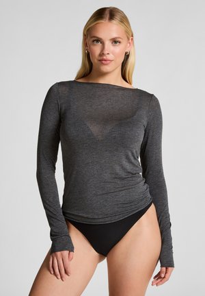 Jonge vrouw in een strakke, lange mouwen donkergrijze top en zwarte hoog uitgesneden onderkant, staande tegen een eenvoudige lichte achtergrond.