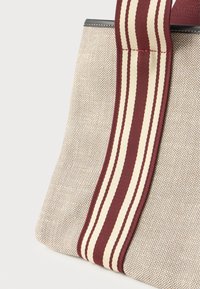 Μπεζ υφασμάτινη τσάντα με ευρείες χειρολαβές σε μαρόν και κρέμ ρίγες, λεπτομερές ράψιμο και μαύρη λεπτομέρεια κατά μήκος της επάνω άκρης.