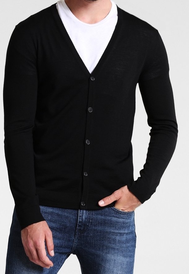 Homme portant un cardigan noir boutonné par-dessus un t-shirt blanc avec un jean bleu, main dans la poche, sur un fond clair uni.