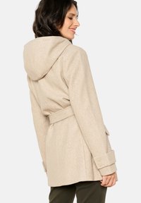 Manteau en laine beige avec capuche, taille ceinturée et poignets à boutons. Présente une texture lisse et une coupe ajustée, adapté pour un usage décontracté.