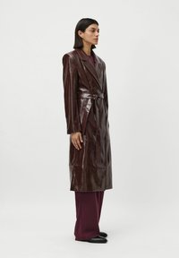 Donna in lungo cappotto di pelle bordeaux scuro con cintura, pantaloni a gamba larga abbinati e scarpe nere, in posizione laterale su sfondo bianco.