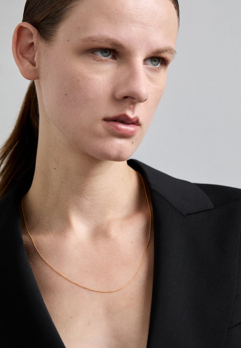 Zlatna verižica, nosena nad črnim blazerjem, z nežnim dizajnom, gladko, sijočo teksturo in subtilno dolžino.
