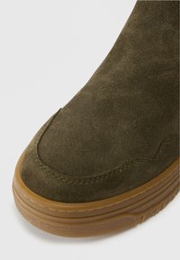 Zelená semišová bota s zaoblenou špičkou, gumovou podrážkou a viditelnými švy podél okraje. Hladký povrch a jemný vzhled.