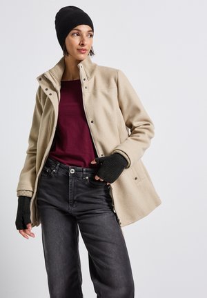 Cappotto in pile beige con colletto alto e bottoni automatici, indossato sopra un top bordeaux e jeans neri. Guanti senza dita in maglia nera e berretto.