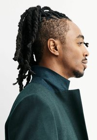Donkergroene jas met een hoge kraag, gemaakt van een gladde stof. De persoon heeft lange, getextureerde dreadlocks. Zijaanzicht.