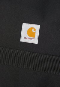 Tissu noir avec une finition texturée, présentant une étiquette blanche avec un logo orange et le texte "carhartt" en bas.
