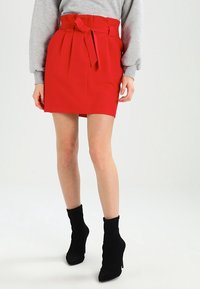 Žena oblečená v červené mini sukni s vysokým pasem, se svázaným páskem, šedém svetru a černých vysokých podpatcích kotníkových bot, stojící na bílé podlaze.