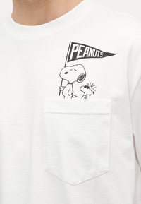 Ο Snoopy κρατά μια σημαία με την ένδειξη "PEANUTS" δίπλα στον Woodstock που φορά γυαλιά ηλίου, εκτυπωμένα πάνω από την αριστερή τσέπη στο μπροστινό μέρος ενός λευκού πουκάμισου.