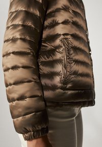 Bruine gewatteerde jas met een gequilt ontwerp, ritsopening aan de voorkant en zijzak. Gemaakt van glad, glanzend materiaal met ribgebreide manchetten.