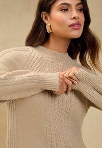 Beige kötött pulóver, melynek elülső részén bordás kötés található, bordázott mandzsettákkal és kerek nyakkivágással, puha tapintással. Elegáns arany fülbevalók vannak viselve.