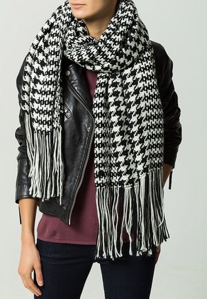 Femme portant une veste en cuir noire, un haut bordeaux et une grande écharpe en pied-de-poule noir et blanc à longues franges.