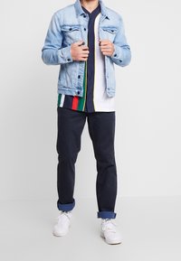 Ljusblå jeansjacka med krage och framfickor. Under den en flerfärgad tröja med horisontella ränder och marinblå byxor. Vita sneakers.