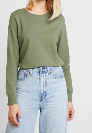 Žena v olivově zeleném tričku s dlouhým rukávem a kulatým výstřihem, zastrčeném do světle modrých džín s vysokým pasem, na neutrálním pozadí.