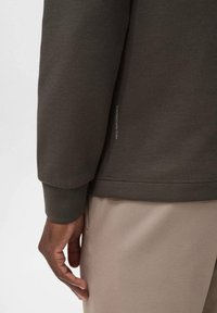 Sudadera verde oscuro con puños y dobladillo acanalados, que presenta un detalle de etiqueta lateral. Textura suave; pantalones caqui visibles abajo.