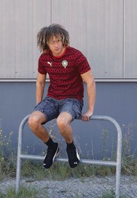 Rød mønstrede sportstrøje med grøn emblem, grå shorts og sorte sneakers. Individuel siddende på et metalgelænder i en udendørs indstilling.