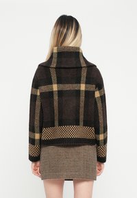 Brauner und schwarzer gemusterter Wollpullover mit einem großen Kragen und einem karierten Design. Über einem kurzen karierten Rock getragen.