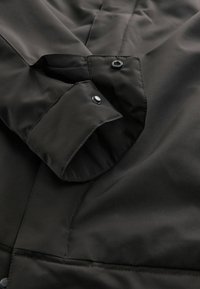 Schwarze Jacke aus glattem, mattem Stoff. Verfügt über eine umgeschlagene Manschette mit Druckknöpfen und sauberen Nähten, die ein strukturiertes Design hervorheben.