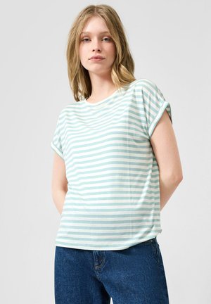 Jeune femme portant une chemise à manches courtes rayée bleu clair et blanc et un jean bleu, debout devant un fond clair uni.