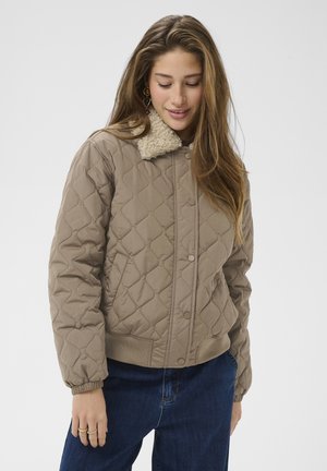 Femme portant une veste beige matelassée avec un col en shearling, un jean bleu et des boucles d’oreilles dorées, regardant vers le bas avec un léger sourire.