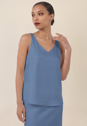 Frau mit dunklem Haar in einem niedrigen Dutt, die ein ärmelloses blaues V-Ausschnitt-Oberteil und einen passenden Rock trägt, posiert vor einem schlichten beigen Hintergrund.