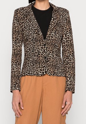 Femme portant un blazer ajusté à motif léopard sur un haut noir et un pantalon marron clair, debout avec les bras détendus le long du corps.