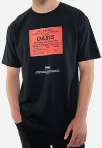 Black cotton t-shirt featuring a large red graphic of an Oasis concert ticket from 1994, text printed in white and red. Textured print detail.
