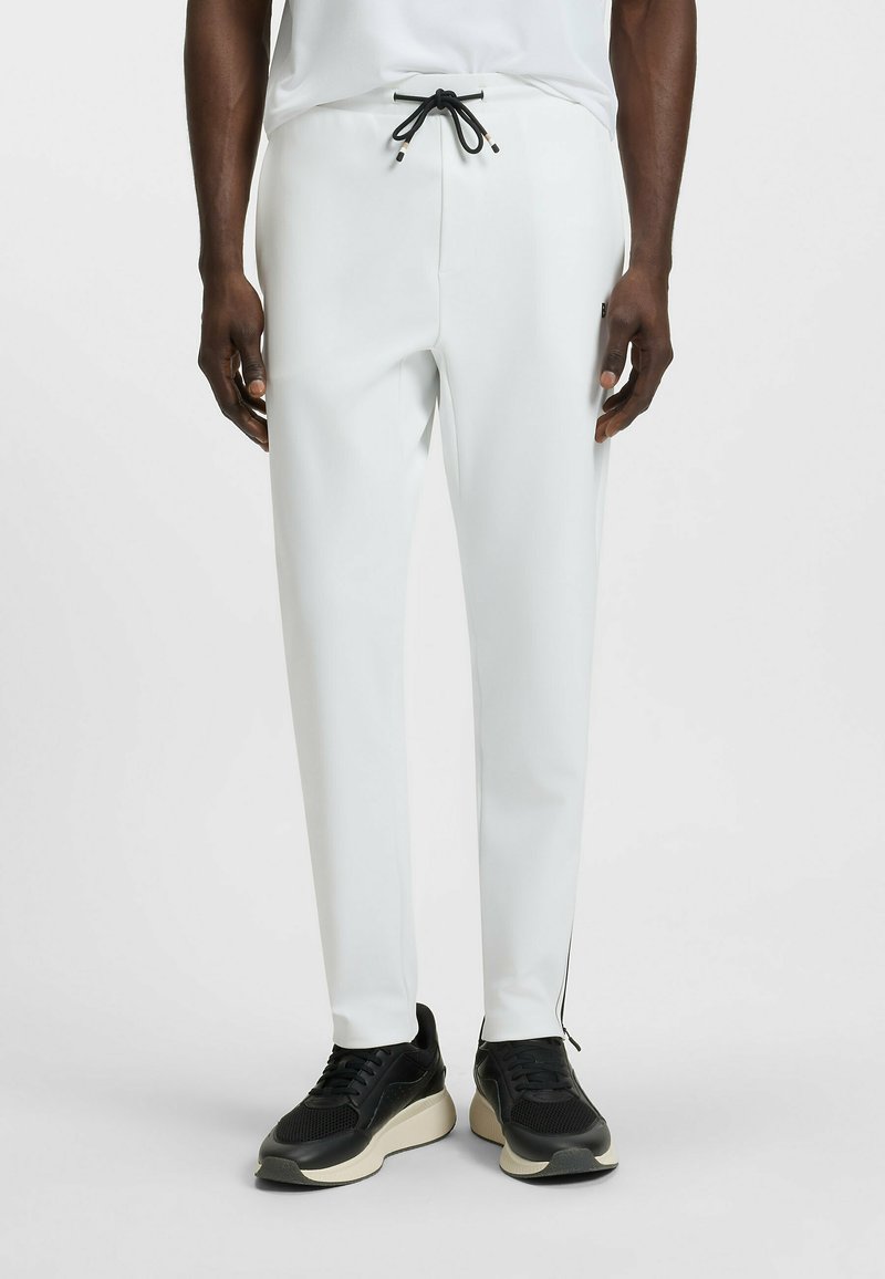 Witte, slim-fit broek met een elastische tailleband en zwarte koord; heeft zijzakken met rits en contrasterende naden. Gepaard met zwarte sneakers.