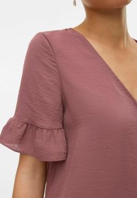 Blusa a maniche corte in malva con scollatura a V, tessuto strutturato e maniche frillate. Focus sui dettagli del collo e delle maniche.