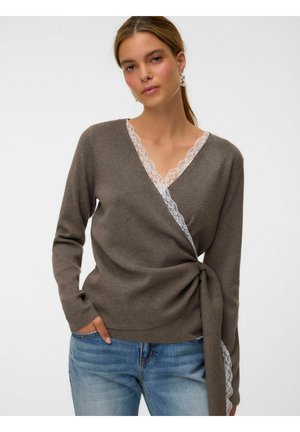 Femme portant un pull cache-cœur marron avec bordure en dentelle blanche, jean bleu et boucles d'oreilles argentées, debout avec une main dans la poche.
