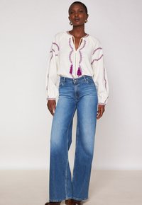 Eine Frau trägt eine weißen Langarmbluse mit lila Stickereien und Quasten, kombiniert mit hochgeschnittenen, weit geschnittenen blauen Jeans und braunen Schuhen.