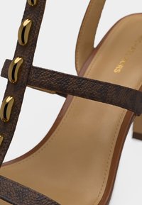 Καφέ λουρί σανδαλιού με υφή, διακοσμημένο με χρυσά οβάλ καρφιά και ταμπά επενδεδυμένη εσωτερική σόλα, με τακούνι block, κοντινή προβολή.