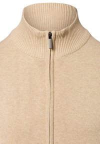 Beige Reißverschluss-Pullover mit hohem Rippkragen und weicher Textur. Verfügt über einen metallischen Reißverschluss auf der Vorderseite und besteht aus Strickstoff.