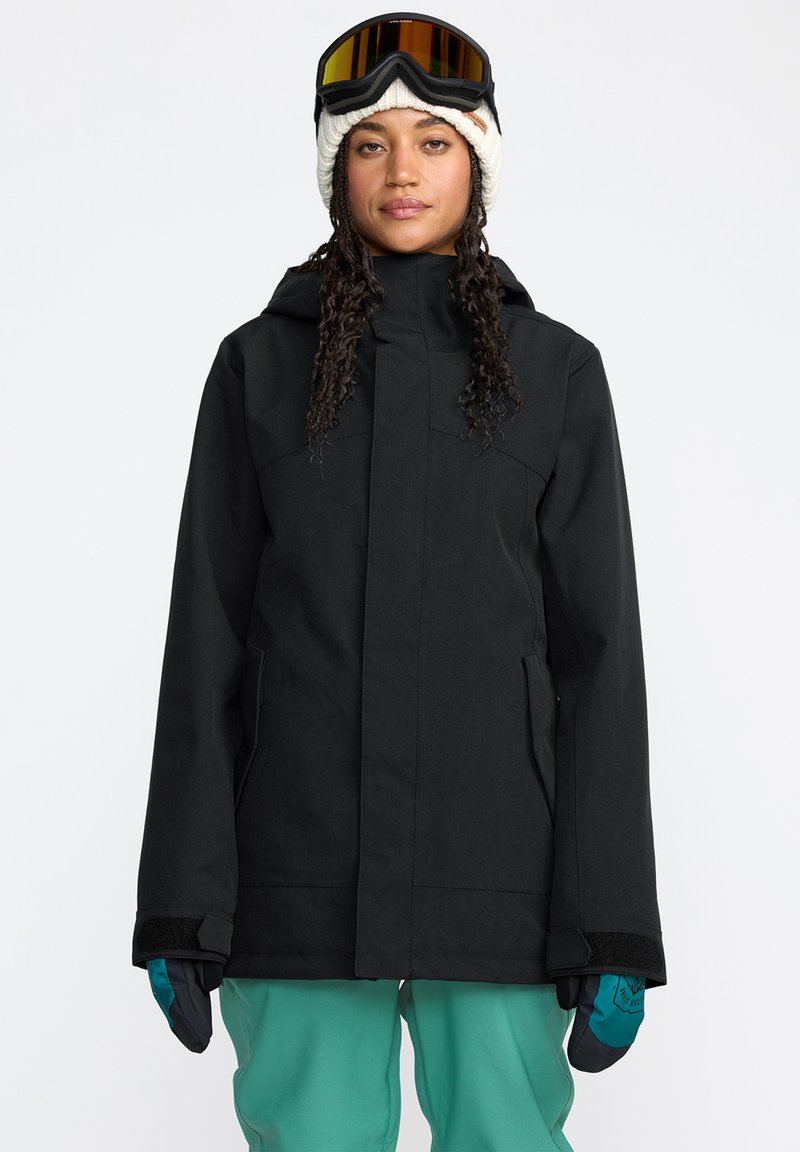 Schwarze Skijacke mit Kapuze, Reißverschluss und Seitentaschen. Getragen mit türkisfarbenen Skihosen und schwarzen Handschuhen, dazu eine gestrickte weiße Mütze und eine Skibrille.