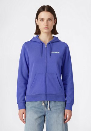 Młoda kobieta o prostych brązowych włosach ma na sobie niebieską bluzę na zamek z logo "CHAMPION" oraz jasnoniebieskie dżinsy, stoi na tle białego tła.