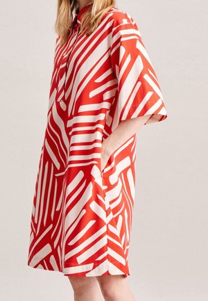 Femme portant une robe ample à manches courtes avec un motif rouge et blanc, col, main dans la poche latérale, sur un fond clair uni.