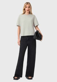 Femme aux cheveux blonds portant un t-shirt ample gris clair, un jean large noir, des sandales noires, et portant un sac à main en cuir noir.