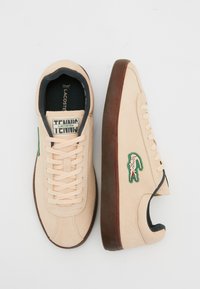 Zapatillas de ante beige con suela de goma marrón, que presentan cordones crema, un logo bordado en verde y un detalle negro alrededor del collar.