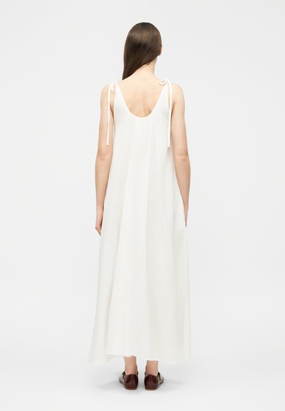 Γυναίκα με μακριά καστανά μαλλιά, φοράει μια λευκή μάξι αμάνικη φόρεμα με κορδόνια και στέκεται γυρισμένη προς τα πίσω, συνδυασμένη με σκούρα καφέ παπούτσια.