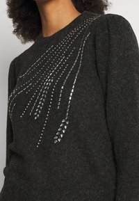 Dunkelgrauer Strickpullover mit einem Perlenmuster und schimmernden silbernen Akzenten, die über eine Schulter verlaufen. Strukturiertes Material.