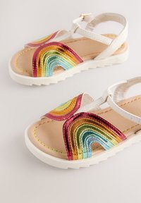 Sandales blanches avec motifs métalliques arc-en-ciel en forme de vagues, dotées d'une semelle plate et d'une bride à la cheville. Texture lisse semblable au cuir avec détails de coutures.