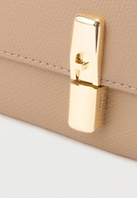 Béžová texturovaná kožená taška v detailu s lesklou zlatou sponou obdélníkového tvaru a jemným prošitím podél okraje.