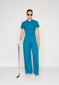 Tyrkysová polo košeľa s golierom, skombinovaná so širokými tyrkysovými nohavicami. Doplnky zahŕňajú biele rukavice a svetlé golfové topánky.