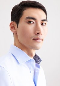 Jonge man met kort donker haar, gekleed in een lichtblauwe overhemdbloes met een gestippelde kraag, kijkt iets naar opzij tegen een eenvoudige witte achtergrond.