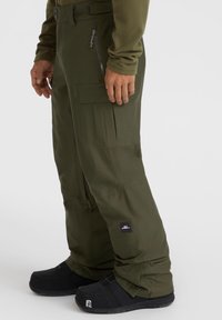 Grüne Schneehosen mit lockerer Passform, ausgestattet mit mehreren Taschen, Reißverschlüssen und einem Stofflabel am Unterschenkel. Kombiniert mit schwarzem Schuhwerk.