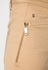 Beiga byxor med sidoficka med dragkedja, guldfärgad hårdvara och en slät tygtextur. Synliga sömmar och en standardmidja.